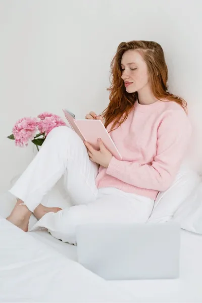 A woman in a pink sweater and white pants sits on a bed with a notebook, next to pink flowers and a laptop.