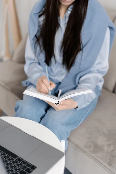 A person sitting on a couch writes in a notebook with a pen, with a laptop open on a nearby table.