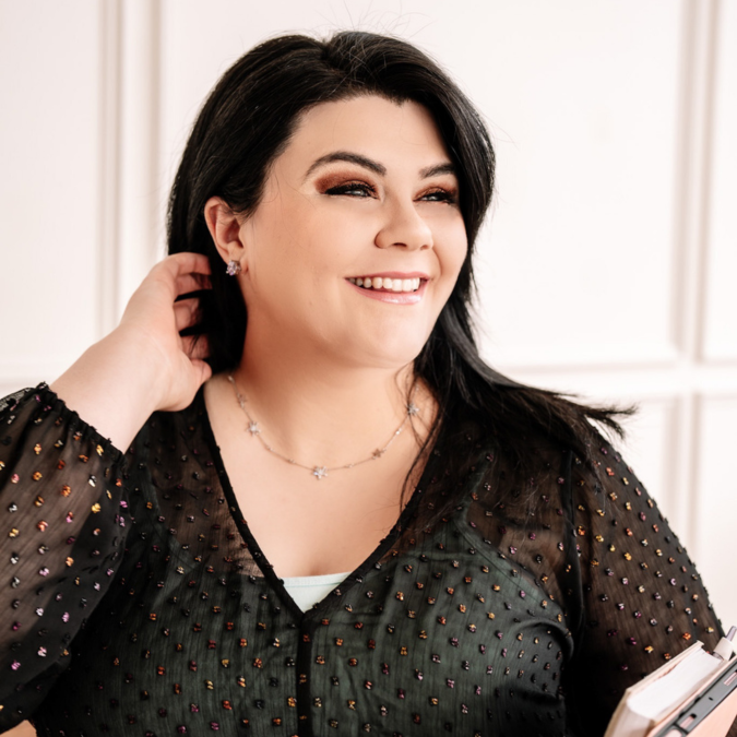 Woman with dark hair in a sheer black top smiles, holding a notebook or planner, standing in a bright room with white paneled walls.
