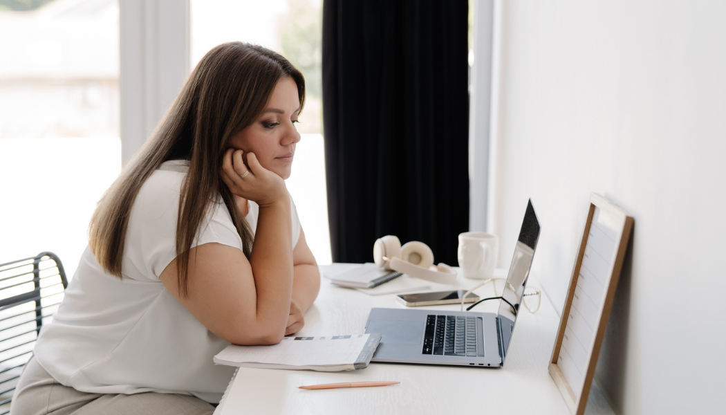 Woman sitting at a desk, looking at a laptop screen with her chin in her hand, contemplating how to get traffic to your website without social media. A notebook, pen, clipboard, and coffee mug are on the white desk.