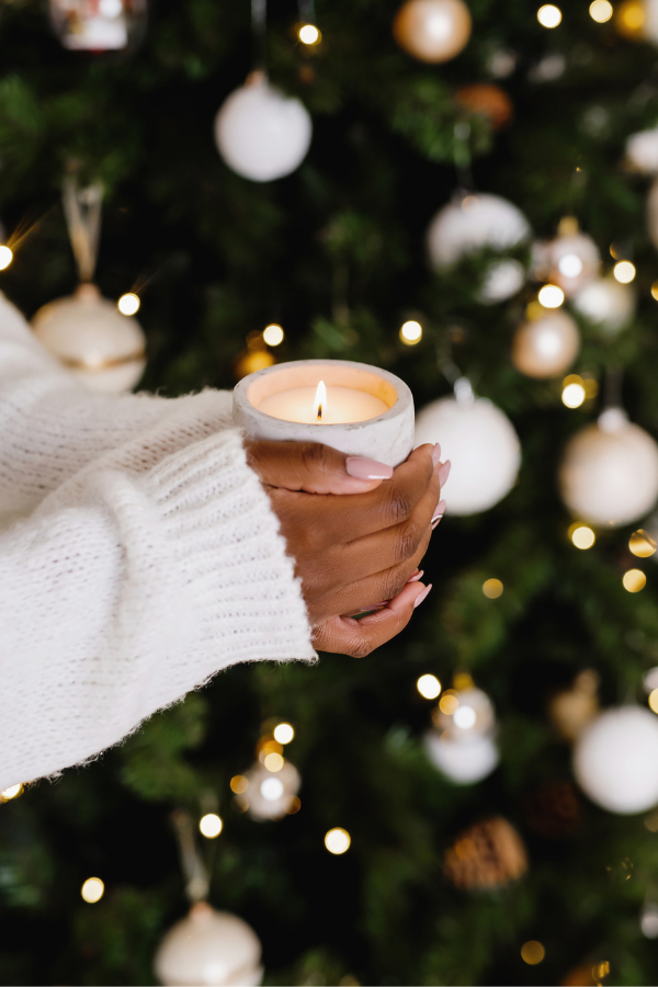 A person in a white sweater holds a lit candle in front of a decorated Christmas tree with white and gold ornaments, capturing the magic that inspires heartfelt holiday copy.